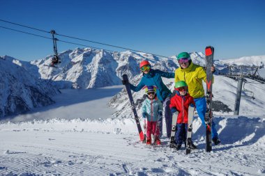 Dört aile ferdi kayak pistinde, ellerinde kayaklarla, nefes kesici dağ manzaralarıyla ve açık mavi gökyüzünün altında duruyorlar.