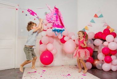 Elinde sopayla zıplayan küçük çocuk pinataya vurmaya hazırlanıyor. Başka bir kız çocuğu da mutlu bir şekilde izliyor.