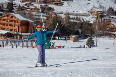 Mavi ceketli ve kırmızı kasklı bir kayakçı, arka planda ahşap pansiyonlar bulunan karlı bir köyün yakınında kayak yapmaktan hoşlanıyor.