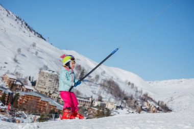 Karlı bir tepede, kışın giyinmiş genç bir kız arka planda bir kayak çekimini, dağları ve Alp tatil köyünü kavrıyor.