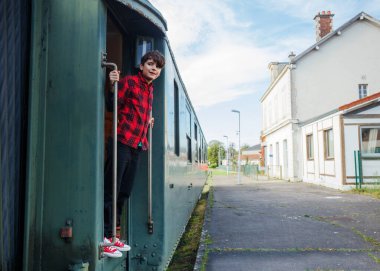 Genç çocuk yeşil vagonun kapısında duruyor, istasyonda ayrılmayı bekliyor, yaz tatili boyunca trenle seyahat etmekten mutlu.