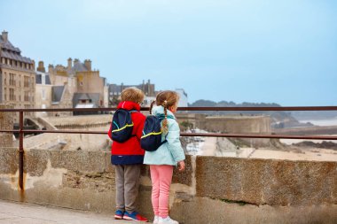 Sırt çantalı iki çocuk, Fransa 'nın Bretagne kentindeki taştan bir bariyerin arkasından Saint-Malo kıyı kasabasına bakıyor.