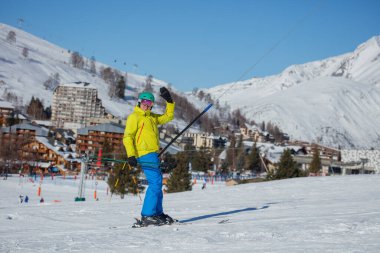 Parlak sarı ceketli bir kayakçı, arka planda binalarla karlı bir dağ otelinde kayak yaparken mutlu bir şekilde el sallıyor.