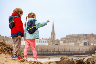 Sırt çantalı iki genç kayalık bir arazide dikilip arka planda Saint-Malo, Bretagne, Fransa 'ya bakıyorlar.