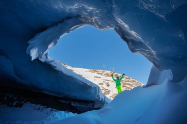 Renkli bir genç kayakçı kar kaplı bir buz mağarasının girişinde duruyor.