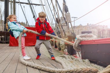 Bir gemi güvertesinde, kalın ceketli bir çift çocuk kalın bir halatı sürüklemek gibi eğlenceli bir işe girişiyor, ilginç bir turistik cazibe.