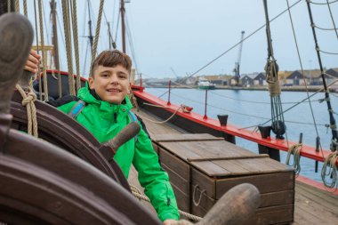 Parlak ceketli neşeli bir çocuk, Saint-Malo, Bretagne, Fransa 'nın rıhtımına yanaşmış tarihi bir geminin direksiyonuna manevra yapıyor.