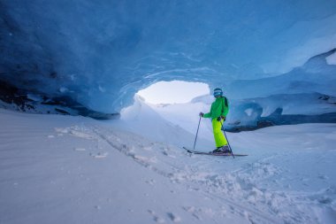 Karlı bir arazide, kayak yapan genç bir çocuk doğal oluşumu takdir etmek için bir buzulun mavi mağarasında durur.