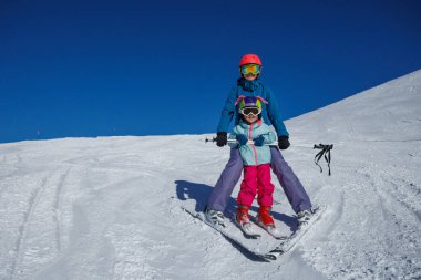 Bir eğitmen genç bir kayakçı kızı hafif bir yamaçta tutuyor, kayak merkezinde fren takozunda süzülüyorlar.