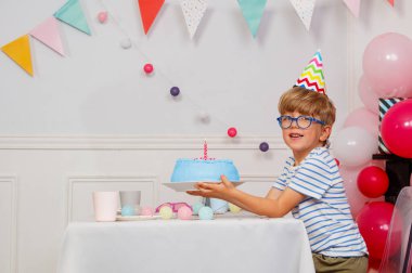 Gülümseyen bir çocuk gözlük takıyor, zigzag desenli bir parti şapkası üzerinde mum olan mavi bir pasta sergiliyor, arka planda çok renkli kiraz kuşu ve balonlar var.