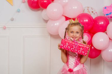 Bir dizi balonun ortasında, genç bir kız elinde hediyesiyle pembe peri kıyafetiyle büyük bir gülümseme sergiliyor.