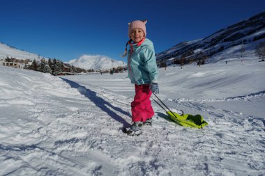 Mavi ceket ve pembe pantolon giymiş genç bir kız, arkasında dağlar olan karlı bir arazide parlak yeşil bir kızak çekiyor.