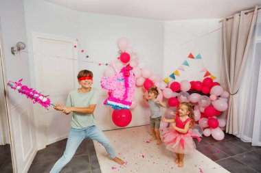 Kutlamanın ortasında, bir genç gözleri bağlı ve elinde sopayla pinataya vurmaya hazırlanıyor, başka bir çocuk da mutlu bir şekilde izliyor.