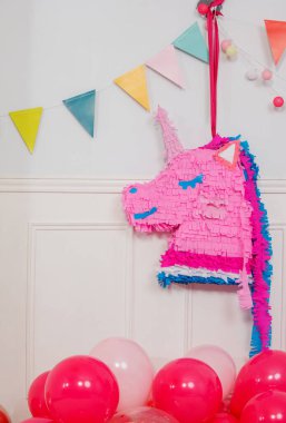 Tek boynuzlu at şeklinde tuhaf pembe bir pinata renkli bayraklar ve küresel ışıklarla süslenmiş bir duvarın yanına yerleştirilir.