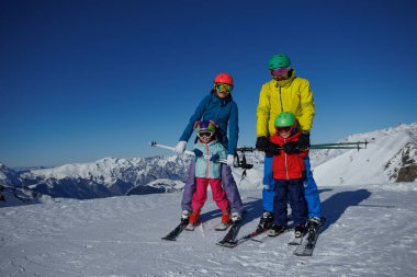 Ebeveynler ve çocuk ikilisi, her biri canlı kask takıyor, arkasında muhteşem karlı tepeler olan kayak yapıyorlar.