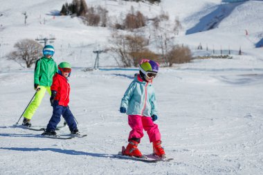Üç çocuk, farklı yaşlarda, renkli kayak kıyafetleri ve kask giymiş kız çocukları, karla kaplı bir yamaçta kayak yapıyorlar.