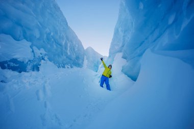 Büyük buz kütleleriyle çevrili dar bir geçitte, sarılı adam zafer işareti sergiliyor.