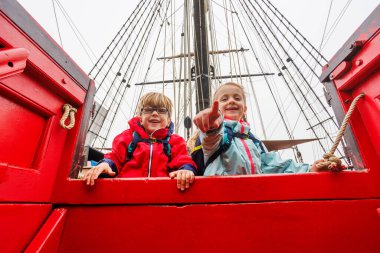Heyecanla, grev ceketli iki çocuk kırmızı bir teknenin kenarından, gemi direğinin altından dışarı bakıyor.