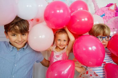 Bir grup çocuk parti atmosferinin tadını çıkarıyorlar. Arka planda canlı balonlar ve şenlik havası var.