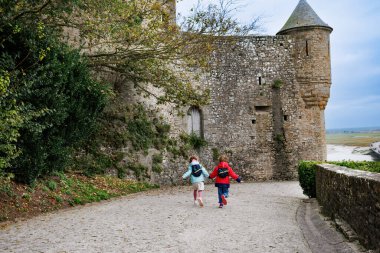 Ortaçağ kalesinin yanındaki kaldırım taşından yolda koşan bir çift çocuk, arka planda resim gibi bir manzaraya sahipler.