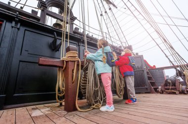Sıcak giyimli bir çocuk ikilisi, turistik bir yerde eski bir yelkenli gemisinin iplerini çekerken resmediliyor.