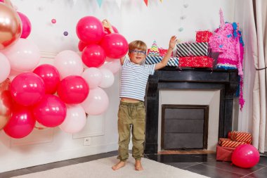 Arkasındaki bacada hediyeler sergilenirken, genç bir çocuk parlak pembe ve beyaz balonların üzerinde durarak kutluyor.