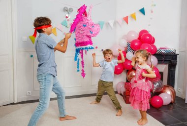 Canlı bir toplantıda, genç bir çocuk gözleri bağlı ve elinde sopayla dekoratif bir pinataya nişan alırken, pembe elbiseli bir kız neşeyle izliyor.