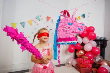 Etrafı şenlikli dekorlarla çevrili pembe kostümlü bir kız doğum günü partisinde tek boynuzlu at pinatasına vurmaya çalışıyor.