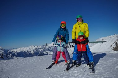 Açık havanın tadını çıkaran iki çocuk ve ebeveynleri dağlık arazide kayak faaliyetinde bulunuyorlar.