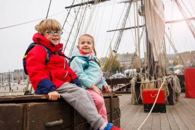Parlak ceketler giyen iki çocuk, bir yelkenli teknesinde ahşap bir sandıkta oturmuş, ziyaretlerinin tadını çıkarıyorlar.