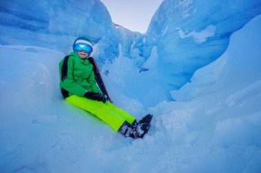 Parlak yeşil ve sarı kayak kıyafetleri içinde gülümseyen genç bir çocuk mavi buz oluşumlarının ortasında oturuyor.