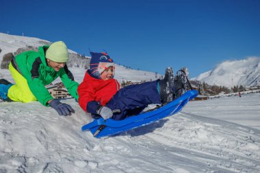 Genç kardeş, kış tatillerinde gülen kardeşiyle birlikte mavi kızağı itiyor.