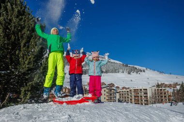 Güneşli tepede, çocuklar zıplar ve havaya kar atarlar, arkalarındaki dağ evinde kayarlar.