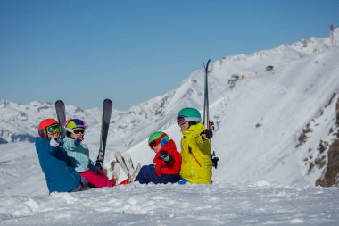 Beyaz dağda oturan dört kişi, anne, baba ve iki çocuk canlı kış kıyafetleriyle karlı ortamın geniş manzarasının tadını çıkarıyorlar.