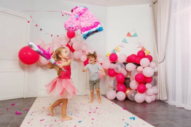 İki oğlan hevesle izlerken, prenses kıyafetli genç bir kız etrafını süsleyen tek boynuzlu at pinatasına vurmaya çalışıyor.