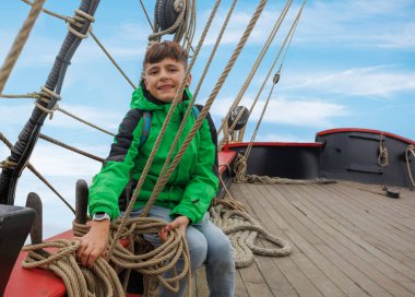 Geminin güvertesinde, turistik bir yerde parlak yeşil bir ceket giyen bir çocuk ipleri kavrarken sırıtıyor, gökyüzü arkasından uzanıyor.