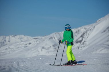 Neon yeşili ceketli genç bir kayakçı yeni bakımlı bir yamacın üzerinde duruyor, direkler soluk mavi bir gökyüzünün altında çıtır çıtır kara dikilmiş.