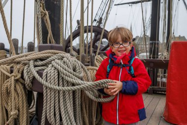 Gözlüklü genç bir çocuk, yelkenli güvertesinde gülümsüyor, parlak kırmızı bir ceket giymiş kalın iplere tutunuyor.