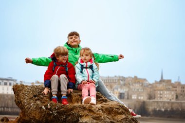 Üç parlak renkli paltolu çocuk deniz kenarında oturuyor ve arkalarında bir şehir manzarası var, Saint-Malo, Bretagne, Fransa