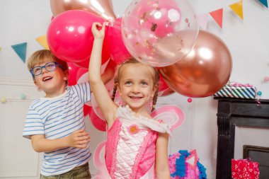 Renkli balonlarla dolu bir odada çizgili tişörtlü ve gözlüklü bir çocuk sırıtırken pembe peri kıyafetli bir kız neşeyle balon tutuyor.