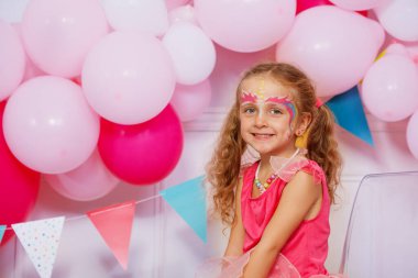 Yüzü fantezi makyajlı kız sırıtırken, pembe balonlar arka planı dolduruyor.