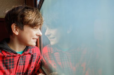 Kırmızı ekose gömlekli genç bir çocuk yaz tatillerinde camdaki yansımasına bakarken gülümsüyor.