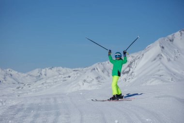 Genç kayakçı eğleniyor. Güneşli bir platoda iki direği de zaferle kaldırıyor. Kar tepeleri gökyüzüne parlıyor.
