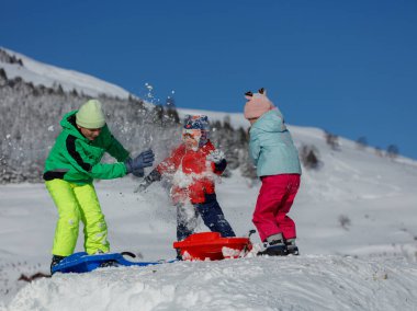 Karla kaplı dağlarla çevrili çocuklar sevinçle birbirlerine mavi gökyüzünün altında kartopu atıyorlar.