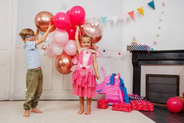 Çizgili bluzlu ve gözlüklü bir çocuk pembe elbiseli bir kızın yanında gülerken görülüyor. İkisi de canlı balonlarla, parti dekorasyonlarıyla çevrili.