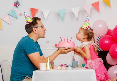 Renkli parti şapkaları takan bir adam ve bir kız, pembe bir doğum günü pastasının üzerindeki mumları üflüyorlar. Etrafı şenlikli süslemeler ve balonlarla çevrili.