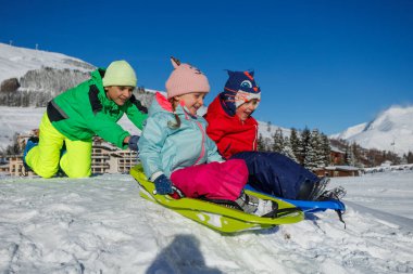 Güneşli bir kış gününde üç neşeli çocuk parlak, renkli ceketleriyle karlı bir yamaçtan aşağı kayıyor.