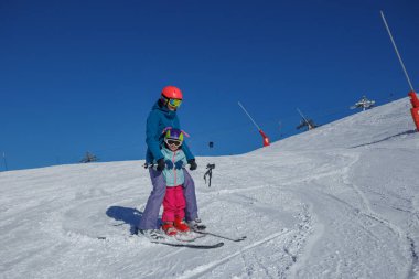 Düz bir yamaçta, bir eğitmen arkada durur ve kızlara kar küreme aracı yapmayı öğretir.