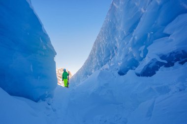 Canlı yeşil dış giyim ve floresan sarı pantolon giymiş bir birey, kayaklarla etkileyici mavi kayalıkların arasında, kaskla poz veriyor.