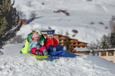 Bir aile, iki çocuk kızaklarda süzülürken yeşil elbiseli bir çocukla karlı bir tepede kızakla kaymanın keyfini çıkarıyor.